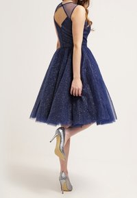 Šatové modré tylové šaty s třpytkami, bez rukávů a V-výstřihem na zádech. Spárováno s lesklými stříbrnými jehlovými podpatky. Plná sukně sahající po polovinu lýtek.