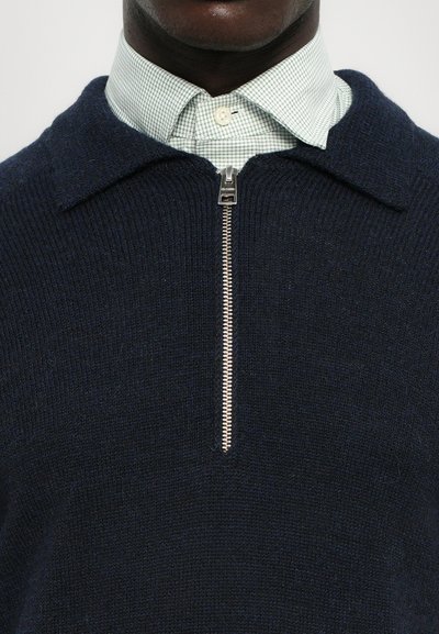 Suéter de lana azul marino con un cuello con cremallera, combinado con una camisa a cuadros en verde claro. Detalle de cremallera plateada notable en el cuello.