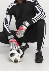 Czarny i biały hoodie adidas z trzema paskami, czerwone i srebrne rękawice bramkarskie, czarne spodnie sportowe oraz piłka nożna w wzory.