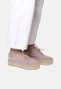 Scarpe espadrillas rosa chiaro con tomaia in stoffa testurizzata, punta rotonda, lacci beige e dettagli in juta intrecciata, abbinate a pantaloni bianchi.