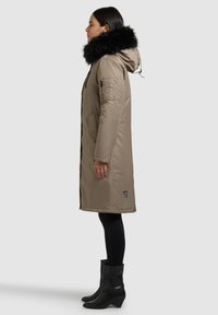 Pikk beež parka musta karusnahaga kapuutsiga. Varustatud tõmblukuga taskute ja logomärgisega. Kandmiseks mustade sukkpükste ja kontsasaabastega.
