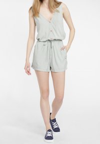 Rompers rayés verts et blancs avec un décolleté en V, une fermeture à boutons à l'avant, une taille élastique et des poches latérales ; associé à des baskets bleu foncé.