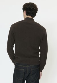 Homme aux cheveux bouclés foncés portant un pull en maille marron foncé et un jean bleu foncé, vu de dos sur un fond clair uni.