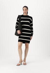 Vestido de malha listrado em preto e branco com mangas longas, corte relaxado e decote careca, combinado com uma mala preta de ombro e sapatos de salto brancos.