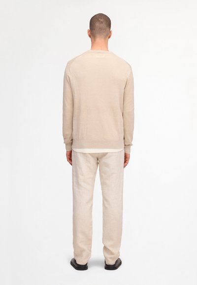 Man wearing beige long-sleeve sweater and matching pants, standing with back facing camera on plain white background.
