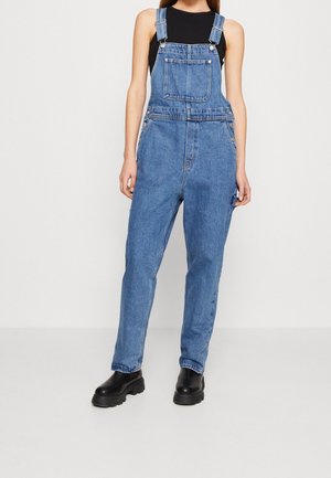 Frau trägt blaue Denim-Latzhose über einem schwarzen ärmellosen Top und schwarzen klobigen Stiefeln, steht vor einem schlichten hellen Hintergrund.