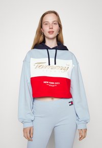 Skosená mikina vo svetlom modrom a červenom prevedení, s námorníckou kapucňou, zlatým logom "Tommy Jeans" a textom "NEW YORK CITY" na červenom panely.