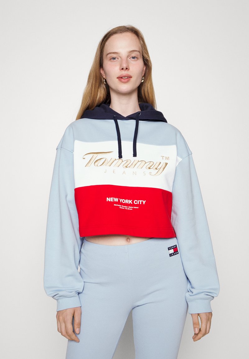 Skosená mikina vo svetlom modrom a červenom prevedení, s námorníckou kapucňou, zlatým logom "Tommy Jeans" a textom "NEW YORK CITY" na červenom panely.