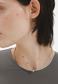 Collar de plata con un diseño circular y suave. Presenta un pequeño acento con detalles en púrpura y dorado en el centro. Textura lisa.