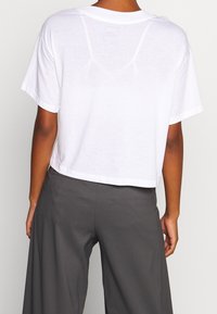 Osoba oblečená v volném bílém crop topu a tmavých širokých kalhotách, zachycena zezadu na jednoduchém pozadí.