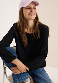 Zwarte lange mouw top met ribbelstructuur, gecombineerd met relaxte blauwe jeans. Een lichtpaarse pet complementeert de casual outfit.