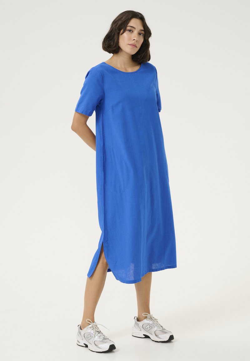 Blaues Leinenkleid mit Rundhalsausschnitt und kurzen Ärmeln, mit Seitenschlitzen. Kombiniert mit grauen Sneakers.