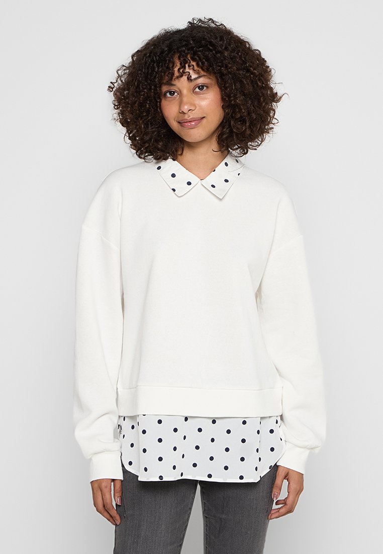 Weißer Pullover mit einem gepunkteten Kragen, ausgestattet mit langen Ärmeln und einem gerippten Saum, über einem weißen Hemd mit schwarzen Punkten getragen.
