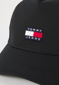 Juodas beisbolo kepurė su stačiakampio formos aplikacija mėlyname fone, baltomis ir raudonomis horizontaliomis juostelėmis ir baltu užrašu "TOMMY JEANS."