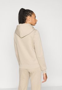 Μπεζ φούτερ με κουκούλα και κορδόνι, ριμπ μανσέτες και τελείωμα. Το μοντέλο φοράει ταιριαστό μπεζ παντελόνι φόρμας, βλέπεται από πίσω, με ελάχιστη υφή.