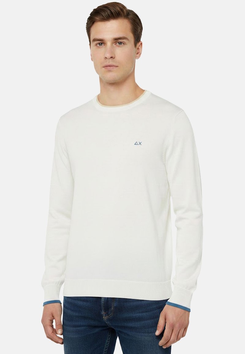 Giovane uomo che indossa un maglione bianco a maniche lunghe con un piccolo logo sul petto e polsini bordati di blu, abbinato a jeans blu, in piedi fermo.