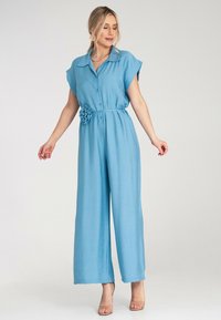 Jasnoniebieski kombinezon z krótkimi rękawami, z przodem zapinanym na guziki, z talią ściągniętą w pasie i dekoracyjnym akcentem w postaci kwiatu w pasie. Szerokie nogawki.