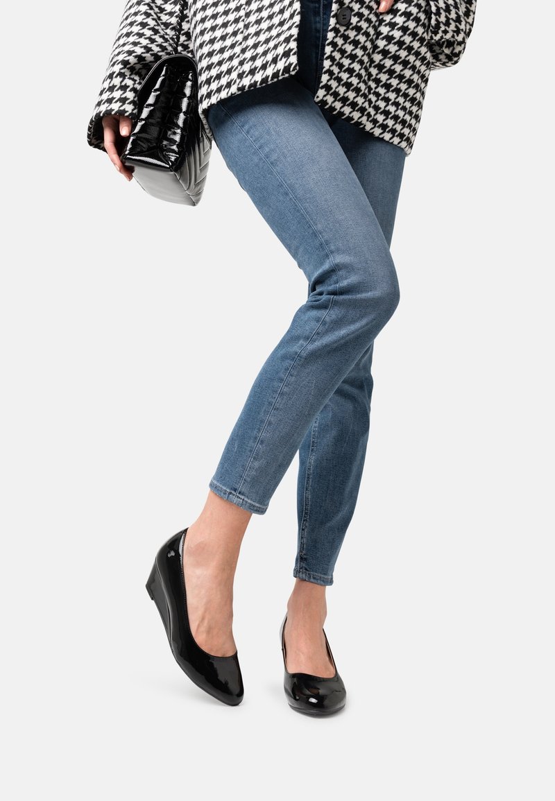 Femme portant un jean slim bleu et des chaussures compensées en cuir verni noir, tenant une pochette matelassée noire, debout avec un pied croisé.
