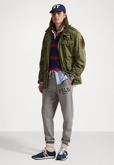 Ung mand iført en olivenfarvet militærjakke, navy og rød stribet rugbytrøje, grå joggingbukser med "Polo" logo, navy sneakers og NY baseballkasket.