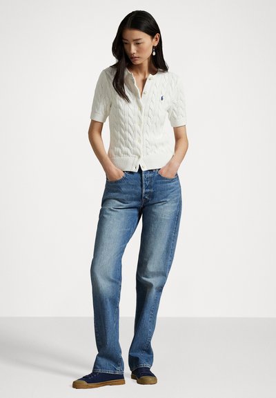Cardigan en tricot à manches courtes de couleur blanche, avec un devant à boutons, porté sur un jean droit bleu et des baskets noires avec une semelle marron.