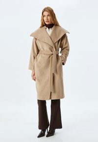 Abrigo envolvente de mezcla de lana beige con gran cuello, cinturón de lazo, mangas largas y bolsillos laterales, combinado con pantalones de pierna ancha oscuros y tacones altos.