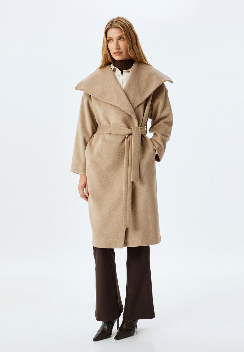 Abrigo envolvente de mezcla de lana beige con gran cuello, cinturón de lazo, mangas largas y bolsillos laterales, combinado con pantalones de pierna ancha oscuros y tacones altos.