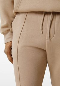 Beżowa bluza z kapturem i dresy wykonane z miękkiego, elastycznego materiału; mają kieszeń kangurzą oraz zakończenia rękawów z regulowanymi sznurkami.