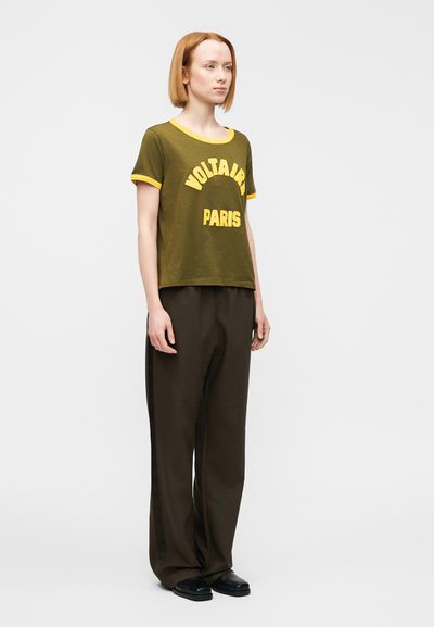 Κοντομάνικη μπλούζα σε ελιά πράσινο με κίτρινο περίγραμμα και τυπωμένο με έντονα γράμματα το "VOLTAIRE PARIS", συνδυασμένη με φαρδιά σκούρα παντελόνια και μαύρα παπούτσια.