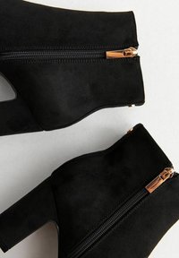 Bottines noires en daim avec un talon bloc, fermeture éclair latérale et un design découpé. Présente des accessoires en métal doré et une texture lisse.