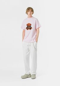 Camiseta oversized de color rosa claro con un gráfico de oso de peluche, combinada con pantalones jogger blancos que tienen bolsillos laterales y un detalle de cadena plateada.