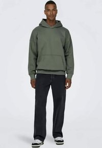 Grüner Pullover-Hoodie mit einer Fronttasche, kombiniert mit dunklen Jeans. Der Hoodie hat eine Kordel und gerippte Bündchen sowie einen Saum. Schwarz-weiße Turnschuhe.
