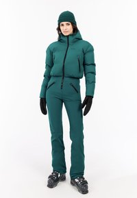 Teal ski-outfit met een puffy jas, aansluitende broek en handschoenen. Voorzien van ritszakken, capuchon en sport-specifieke schoenen.