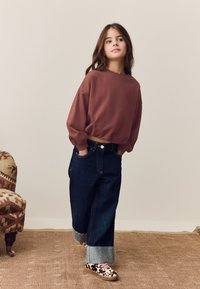 Tummanpunainen lyhyt collegepaita, jossa roikkuvat olkapäät, yhdistetty tummansinisillä farkuilla, joissa on käännetyt lahkeet ja vaaleansinisiä yksityiskohtia; jalassa kuvioidut lenkkarit.