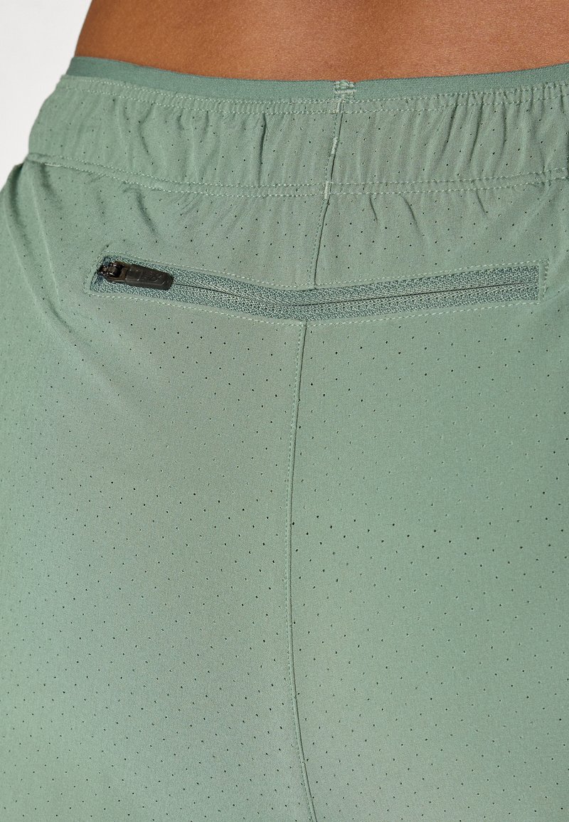 Pantalones cortos deportivos de color verde claro con un tejido texturizado y pequeñas perforaciones. Incluyen un bolsillo con cremallera seguro en la parte trasera de la cinturilla.