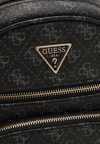 Gros plan sur un sac à dos Guess noir avec un logo triangulaire doré, un motif répété "GS" et deux fermetures éclair dorées horizontales.