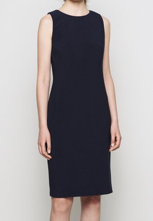 Hihaton, polveen ulottuva istuva laivastonsininen mekko, jonka päällä seisova henkilö yksiväristä vaaleaa taustaa vasten.