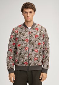 Giacca bomber con un motivo floreale e labbra rosse, in tonalità terrose e un tessuto testurizzato. Polsini a costine e chiusura zip completano il design.