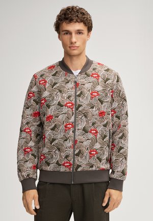 Bomber jacket featuring a floral pattern with red lips, in earthy tones and a textured fabric. Ribbed cuffs and a zip closure complete the design.