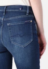 Mørkeblå denimjeans har et tilpasset design, med to baklommer og sydd detaljer. En merket lapp er synlig i midjen.