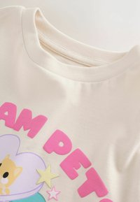 Flødefarvet t-shirtstof med rund halsudskæring. Indeholder pink, hævet tekst og en grafik af en tegneseriefigur med stjerner.