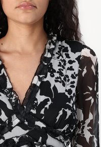 Crna i bijela bluzica s cvjetnim uzorkom, s naboranim izrezom, prozirnom tkaninom i dugim rukavima. Ima detaljno crno šivanje.