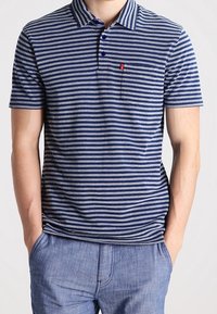 Homme portant un polo à rayures horizontales bleu et gris à manches courtes, avec col et poche poitrine, assorti à un pantalon décontracté bleu clair.