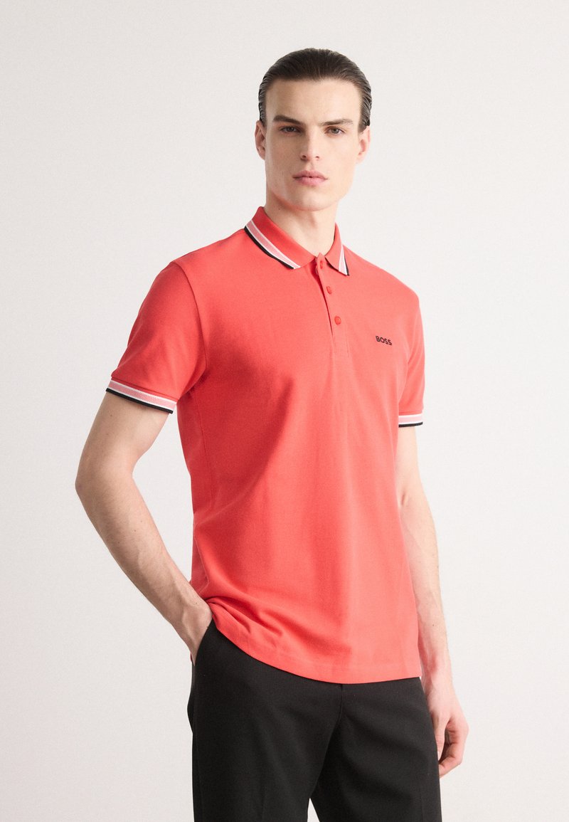 Koral polo-skjorte med svart og hvit stripete krage og ermer, med en subtil logo på brystet, laget av pustende stoff.