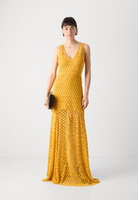 Gul lacedrakt med V-hals, tettsittende silhuett og blomsterdesign. Flyter ned til gulvet med et gjennomsiktig stoff, kombinert med en svart clutch.