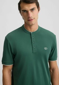 Zelená polo košeľa s okrúhlym golierom, zapínaním na päť gombíkov a kontrastnými bielymi akcentmi na golieri a rukávoch. Obsahuje malé logo.