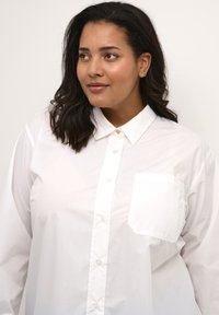 Camisa blanca de botón hecha de una tela ligera, con un cuello clásico, bolsillo frontal y textura suave.