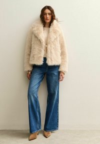 Kvinna som står mot en vägg iförd en beige fuskpälsjacka, blå vida jeans, beige spetsiga klackar och guldsmycken.