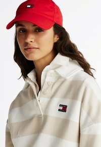Rode pet met een logo aan de voorkant, beige en wit gestreept pullover shirt met kraag en knoopsluiting, voorzien van een klein merkpatch.