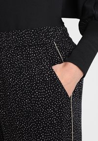 Czarne spodnie z teksturowanej tkaniny, zdobione rozproszonym wzorem białych kropek. Elastyczny pas, boczne kieszenie oraz kontrastowe detale wykończenia.