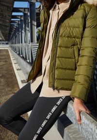 Olivengrøn pufferjakke med pelsforet hætte, beige skjorte med knapper og sorte leggins med hvid tekst på siden. Casual, sporty outfit.